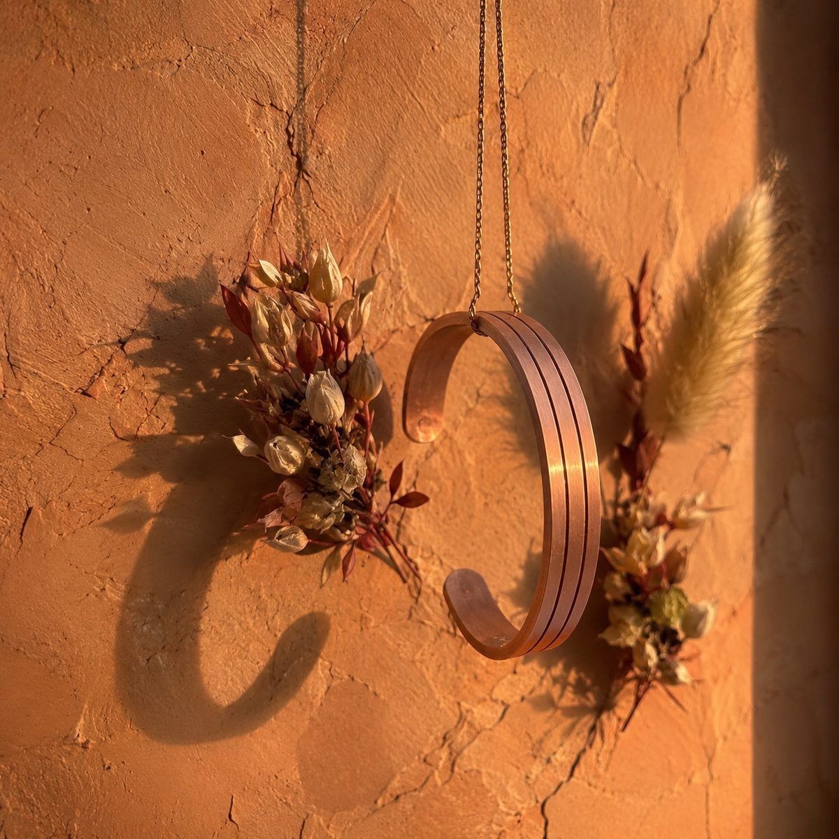 Cuivre Bérénice copper cuff bracelet hanging on a chain against a terracotta wall with dried flowers